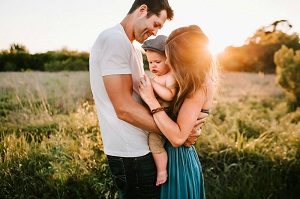 A young couple holding a baby in what looks to be a field, with the sunshine in the background, everyone is very  happy