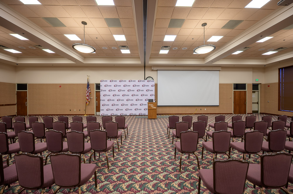 The image is of the Student Center Ballroom. There is a large, white screen on the back wall next to a white backdrop with CSC logos spread across the backdrop. There is a podium in between those two. There are also around 70 purple chairs facing towards the podium.