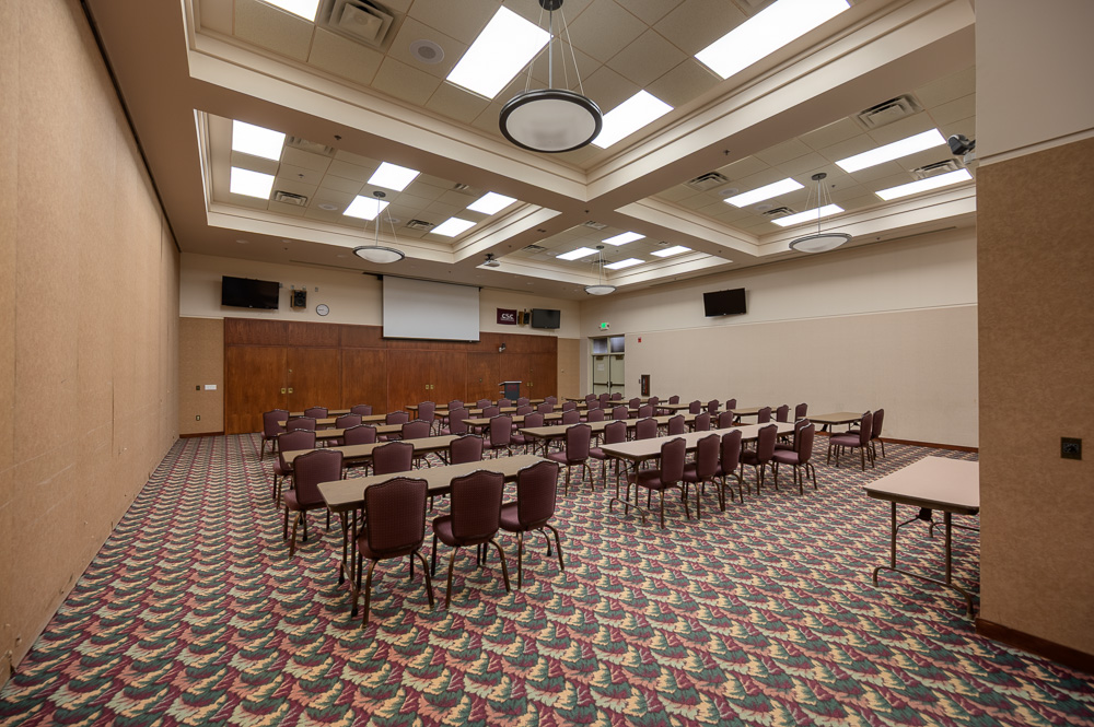 The image is of the Scottsbluff room in the Student Center. The image is of a carpeted room with twenty rectangle tables with three chairs per tables facing the back wall. The back wall has six wooden closed doors and a white screen lowered above them. There are also three TVs and a CSC flag on the top of the walls.