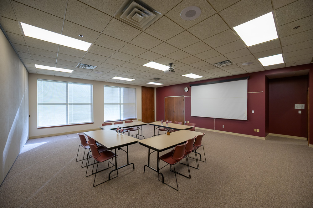 The image is of the Student Center Ponderosa Room. There are two large windows that take up the entirety of the wall to the left of the tables. There are four tables in a square shape with three chairs per tables. The wall to the right of the windows is maroon with two closet doors and a large, white screen lowered against it. To the right of the white screen is the entrance to the room.
