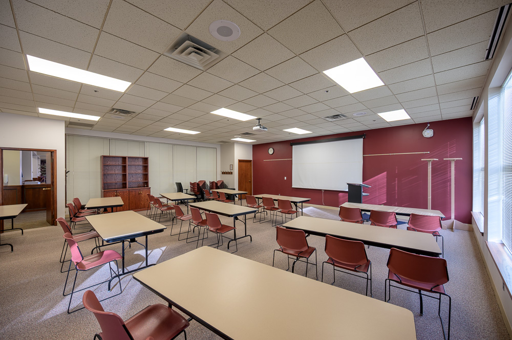 The image is of the Student Center Bordeaux room. There is a large, white screen lowered on the back, maroon wall. There are nine rectangle tables with three chairs each table, facing the screen. There is a podium to the right of the screen and a wooden cupboard against the wall to the left of the tables.