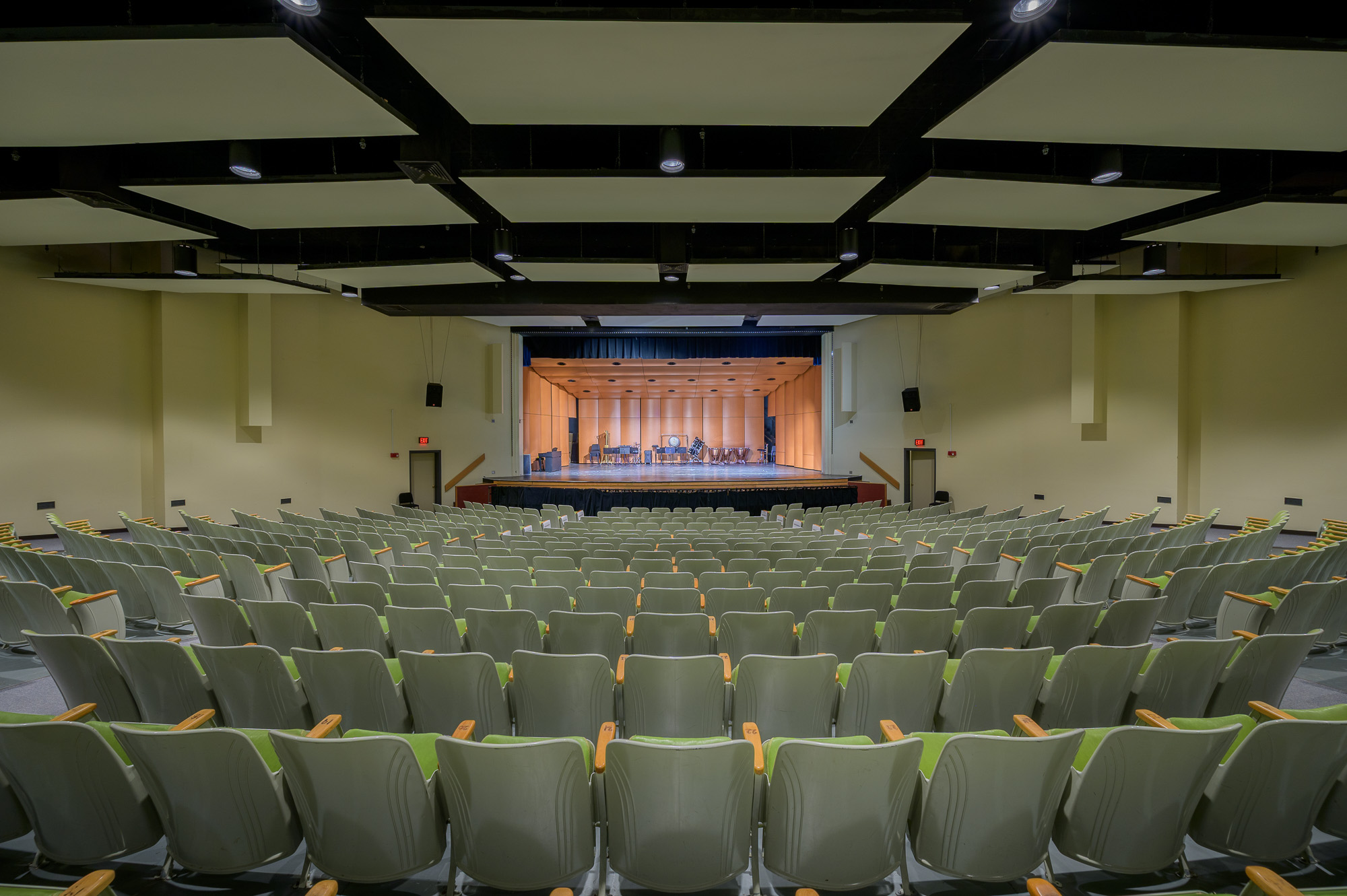 The image of the Memorial Hall Auditorium from the last row of the seats. The very back of the photo is the stage with two exit doors on each side. There are over 10 rows of chairs in the middle and on both sides.