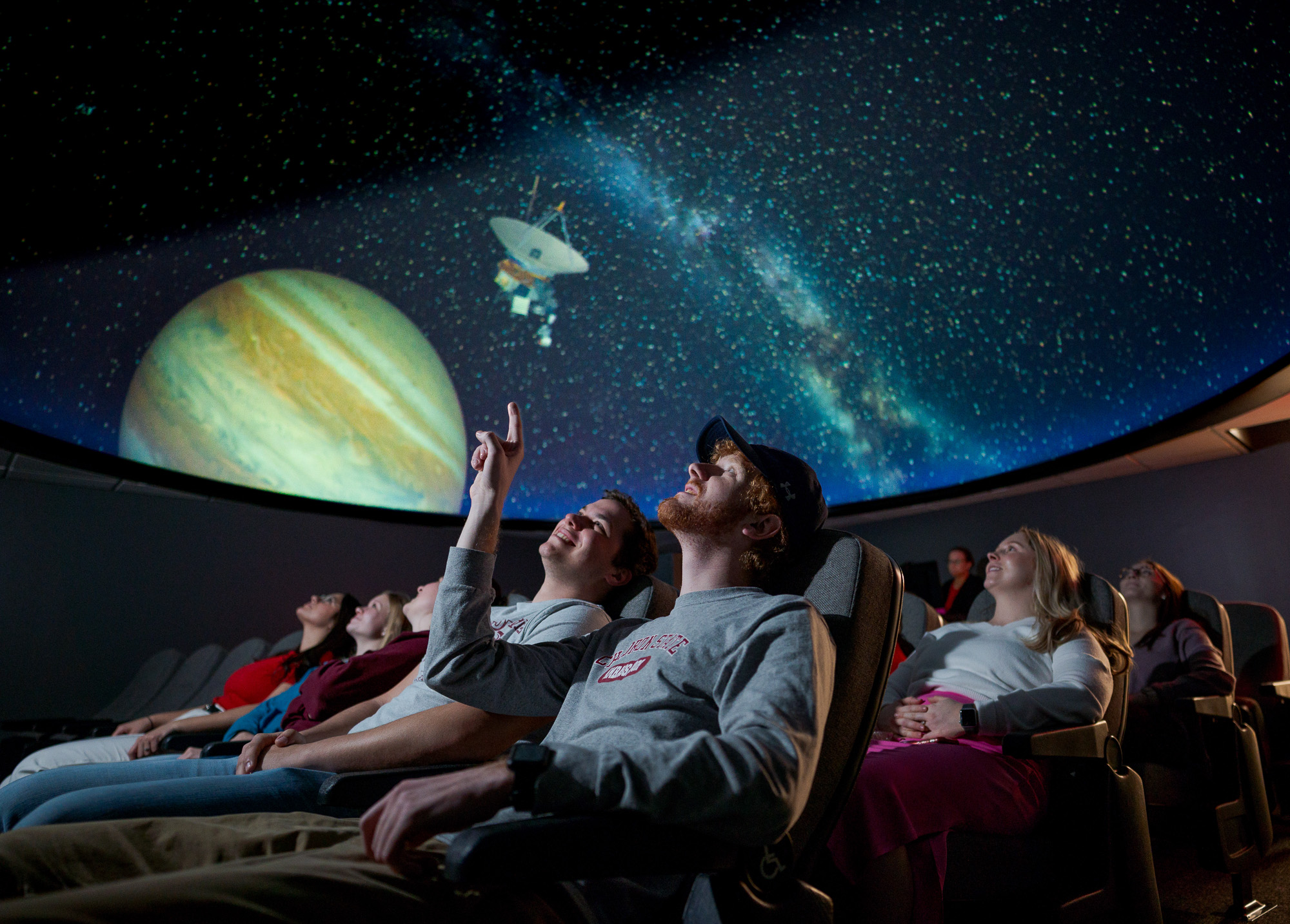 The image shows a bright star filled ceiling with an image of a satellite next to Jupiter. There are people in seats staring up at the ceiling. One student is point at the ceiling.