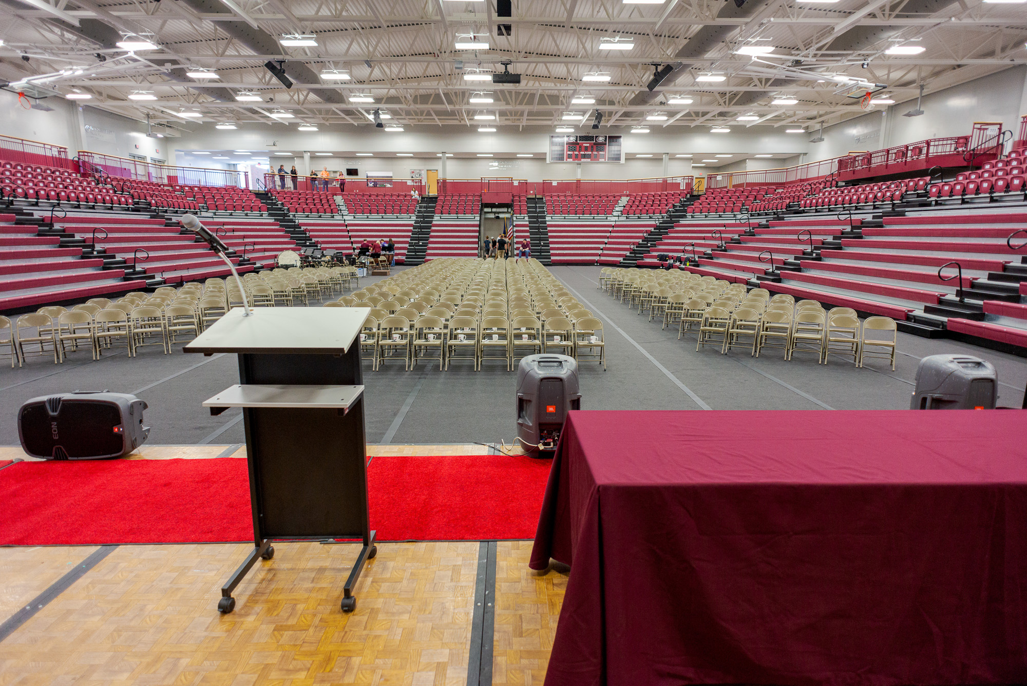 The image is of the Chicoine Center Gym during Commencement. The image shows rows of red bleachers that cover the entire room. In the middle of the bleachers is the actual gym where there are three sections of folded chairs. The front of the image shows a stage with a podium, three speakers, and a table with a maroon tablecloth.