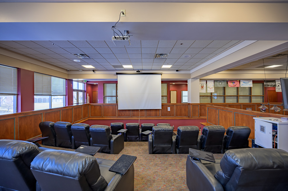 The image is of the Student Center area known as the Pit Theater. The image is of 15 black reclining chairs all facing a large white screen with a projector on the ceiling facing the screen.