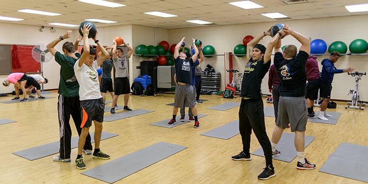 Students in a gym passing weighted balls to each other, with mats on the floor