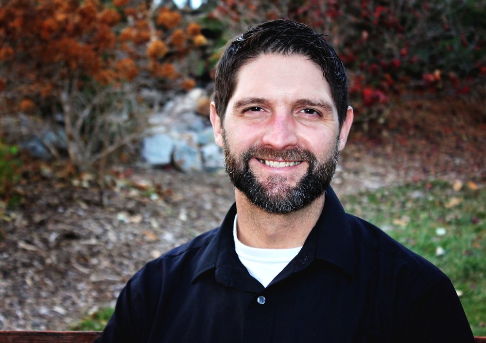 Photo of Keith Buswell smiling in front of foliage.